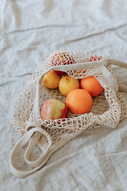 A variety of fresh fruits in a reusable mesh bag placed on a soft linen surface.